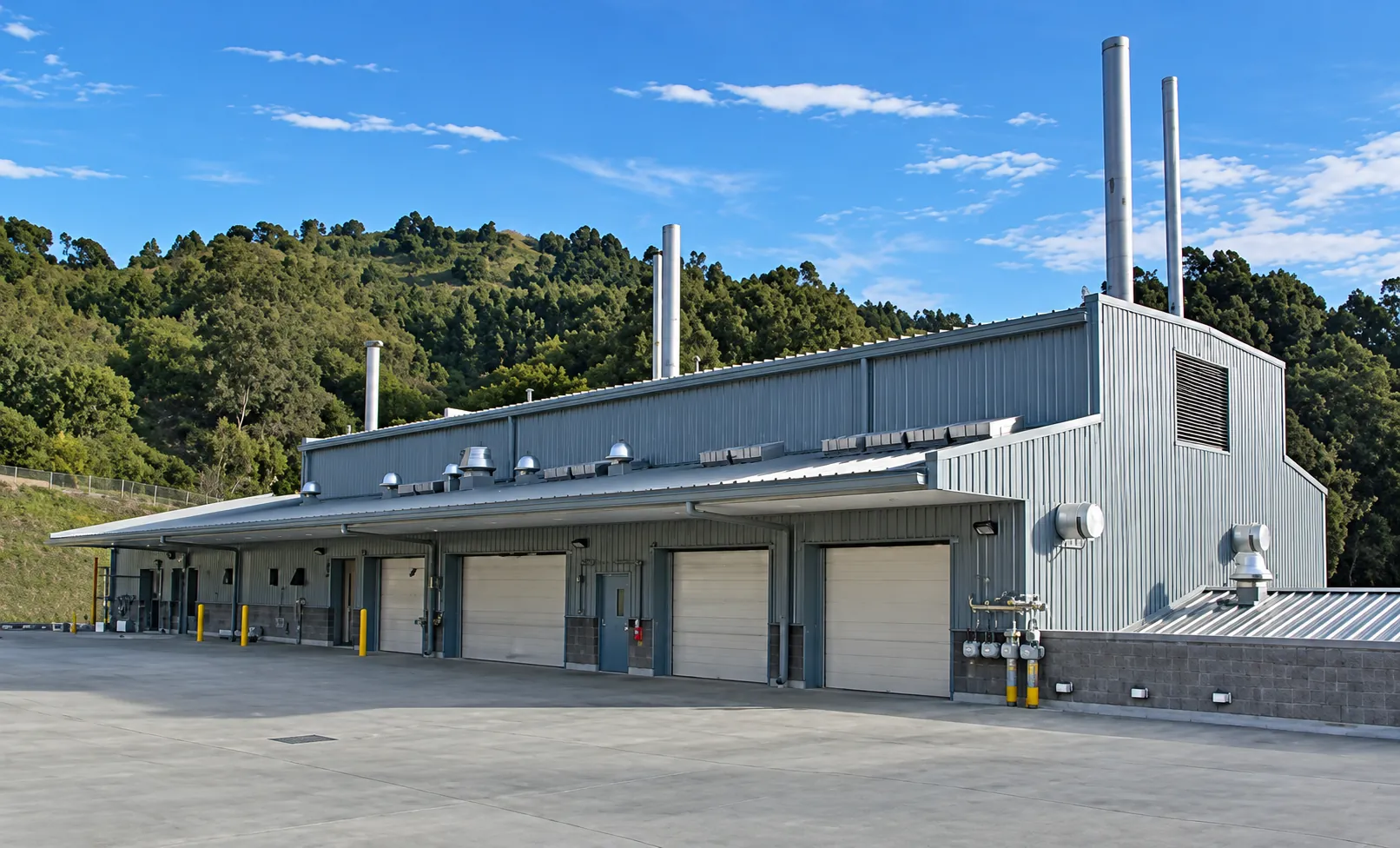 Building 85 Waste Handling Facility at Lawrence Berkeley National Laboratory in Berkeley, California: a metal-clad building with roll-up loading bay doors and exhaust stacks below the wooded Berkeley hills